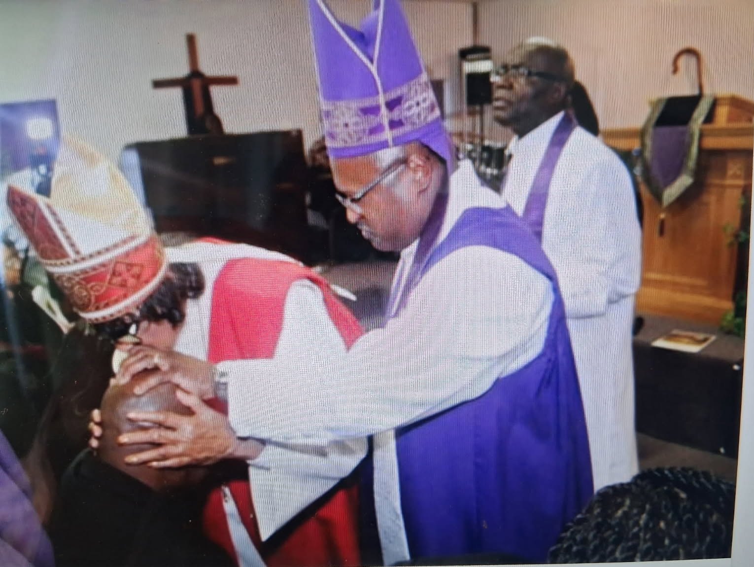 A bishop in a purple robe and mitre laying hands on a kneeling minister in prayer